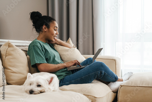 Smiling black woman using laptop while sitting with her dog on sofa