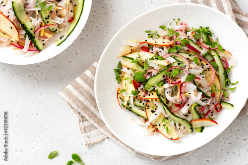 Rice noodle salad with cucumber, apple, onion, sesame seeds, and herbs in a white bowl on a light concrete background top view. Vegan salad Asian style. Vegetarian food.