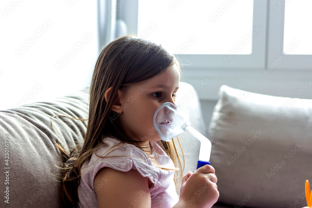 Little girl making inhalation with nebulizer at home. Close-up of girl using asthma inhaler at home. Little girl is suffering from health issues. Child with asthma inhaler at home. Copy space. Little girl making inhalation with nebulizer at home. Close-up of girl using asthma inhaler at home. Little girl is suffering from health issues. Child with asthma inhaler at home. Copy space.