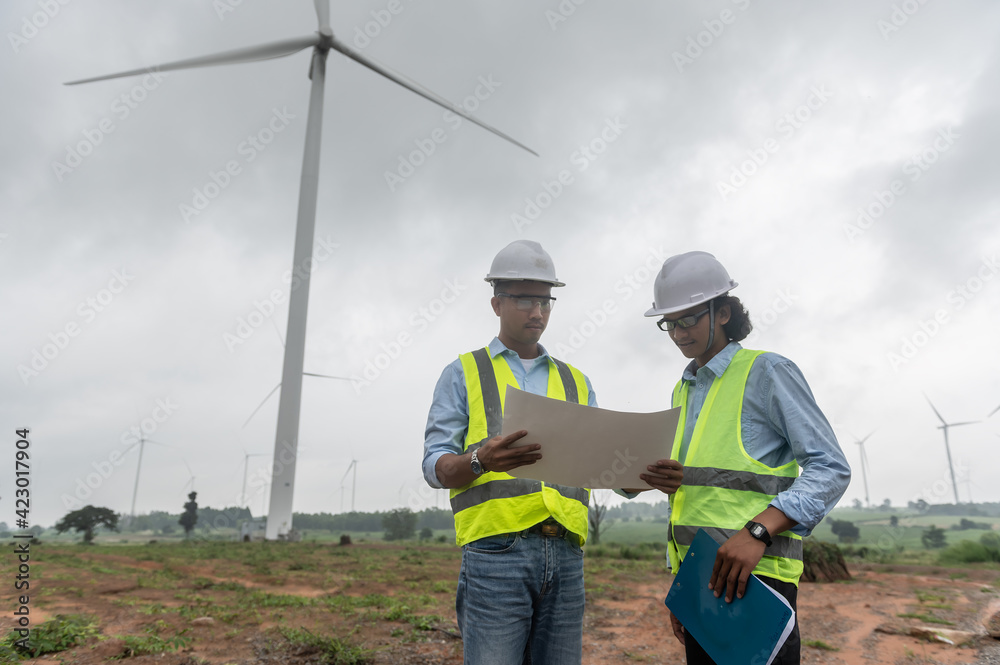 Two engineers working and holding the report at wind turbine farm Power Generator Station on mountain,Thailand people Two engineers working and holding the report at wind turbine farm Power Generator Station on mountain,Thailand people