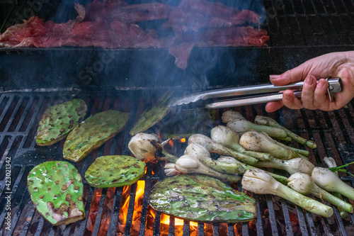 Fotografie front shot of a grill, carne asada, nopales and charcoal grilled cambray onions while one hand is cooking them