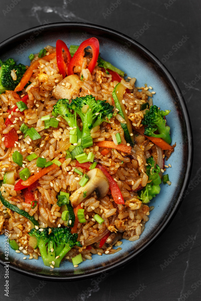 Asian rice with vegetables: zucchini, broccoli, red bel pepper, mushrooms, carrot, spring onion and sesame seeds. Dish isolated in a blue bowl, close-up on a black marble background. Asian cuisine.