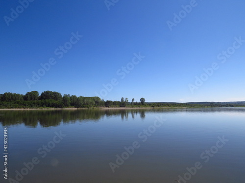 Fototapeta Naklejka Na Ścianę i Meble -  Quiet surface of the river. Beautiful cloudless warm summer day. Trees on the island in the background