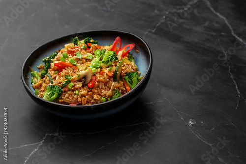 Asian rice with vegetables: zucchini, broccoli, red bel pepper, mushrooms, carrot, spring onion and sesame seeds. Dish isolated in a blue bowl, close-up on a black marble background. Asian cuisine.