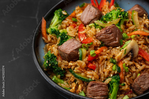 Asian rice with teriyaki beef and vegetables: zucchini, broccoli, pepper, mushrooms, carrot, onion and sesame seeds. Dish isolated in a blue bowl, close-up on a black marble background. Asian cuisine.