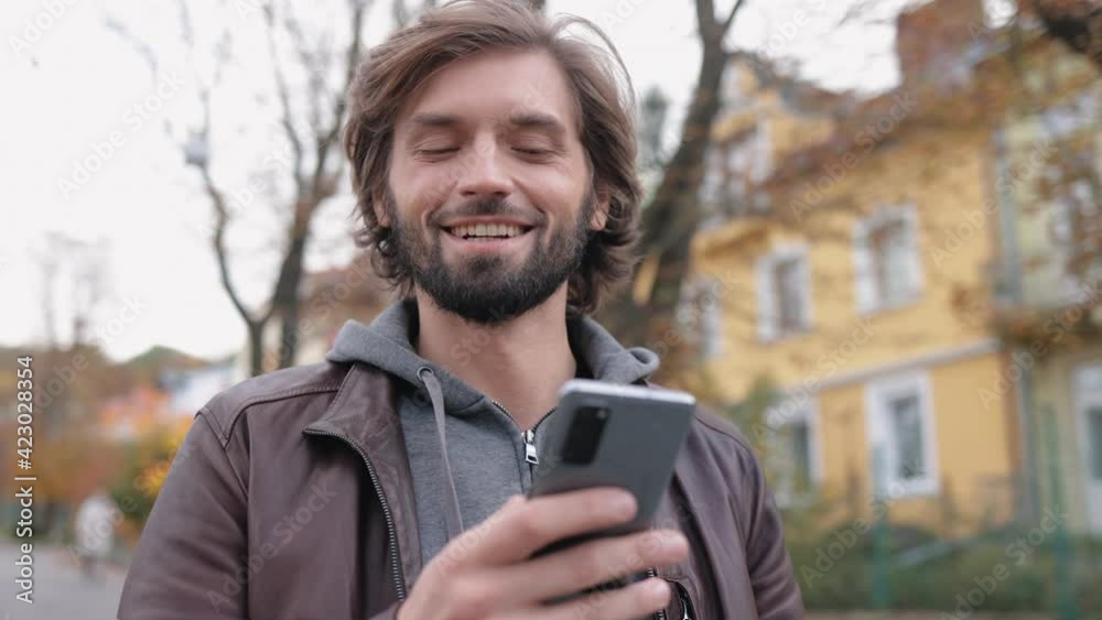 Handsome Bearded Man Chatting on Social Media while Walking on the Street. Positive Man wearing Casual Clothes Typing on Smartphone Outdoors. Having a Good Mood enjoys Strolling. Mobile Device. Apps. Handsome Bearded Man Chatting on Social Media while Walking on the Street. Positive Man wearing Casual Clothes Typing on Smartphone Outdoors. Having a Good Mood enjoys Strolling. Mobile Device. Apps.