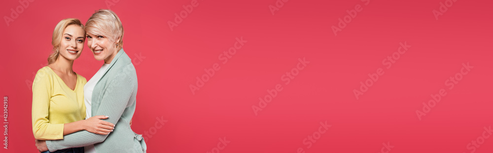 cheerful, mature woman embracing with adult daughter isolated on pink, banner cheerful, mature woman embracing with adult daughter isolated on pink, banner