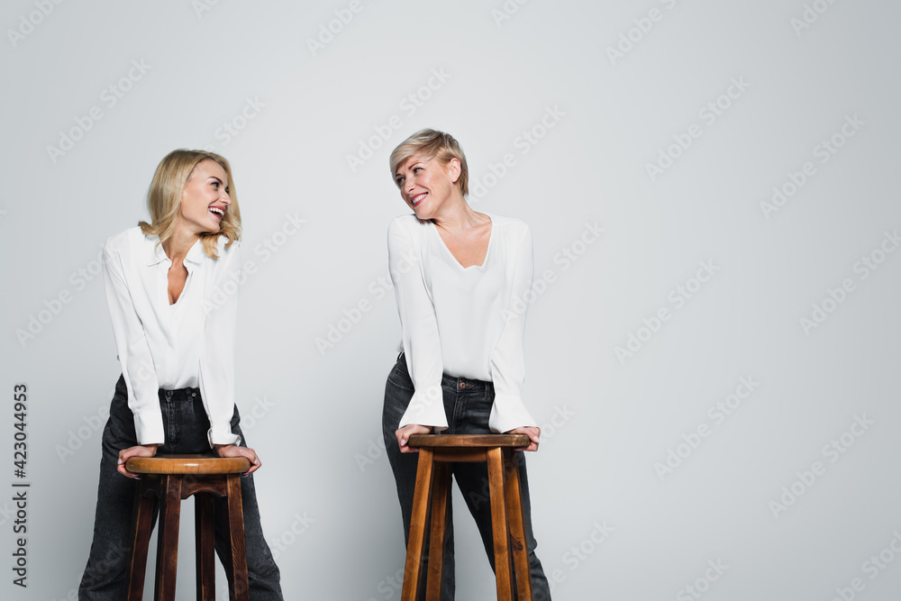 Obraz premium joyful mother and daughter smiling at each other while posing near high chairs on grey
