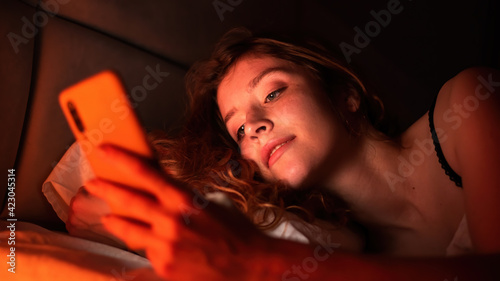 Young girl with smartphone in a bed. Red illumination