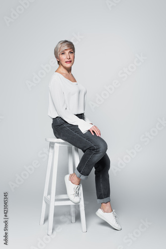 middle aged woman in jeans and white blouse sitting on high chair on grey photo