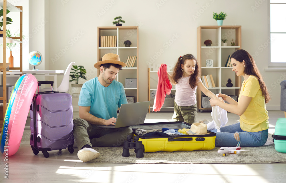 Happy family of three going to travel. Father choosing tour, buying ticket or reserving hotel online using laptop. Mother with preschool daughter packing clothes in suitcase luggage bag. Home interior Happy family of three going to travel. Father choosing tour, buying ticket or reserving hotel online using laptop. Mother with preschool daughter packing clothes in suitcase luggage bag. Home interior