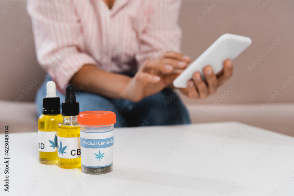 bottles with cbd and medical cannabis lettering near african american woman using smartphone on blurred background