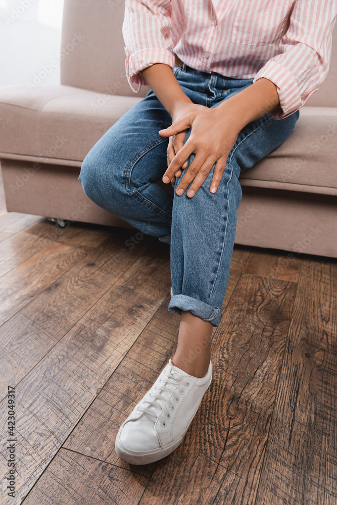 partial view of african american woman suffering from knee pain while sitting on couch