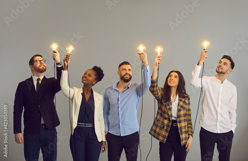 Group of happy creative young diverse business professionals holding glowing light bulbs standing on gray studio background. Innovative thinking, finding solution, people developing own idea concept