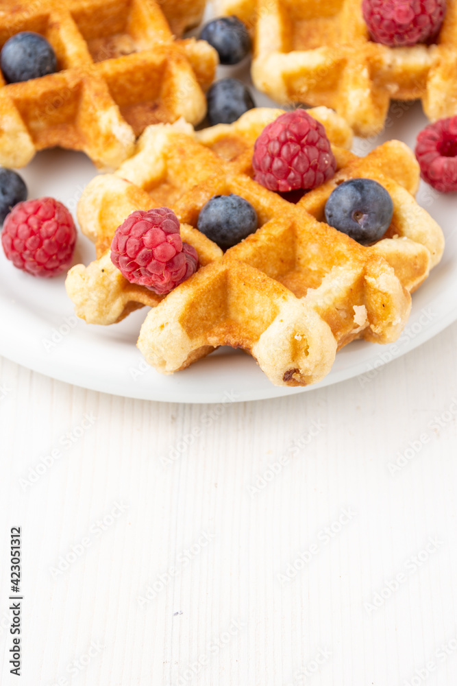 Top view of waffles with raspberries and blueberries, on plate, selective focus, on white table, in vertical, with copy space Top view of waffles with raspberries and blueberries, on plate, selective focus, on white table, in vertical, with copy space