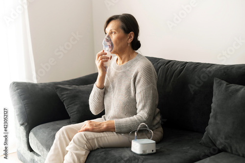 Tela Retirement age woman using inhaler for flu and cold treatment sitting on the sofa at home, senior older female cover face with an oxygen mask