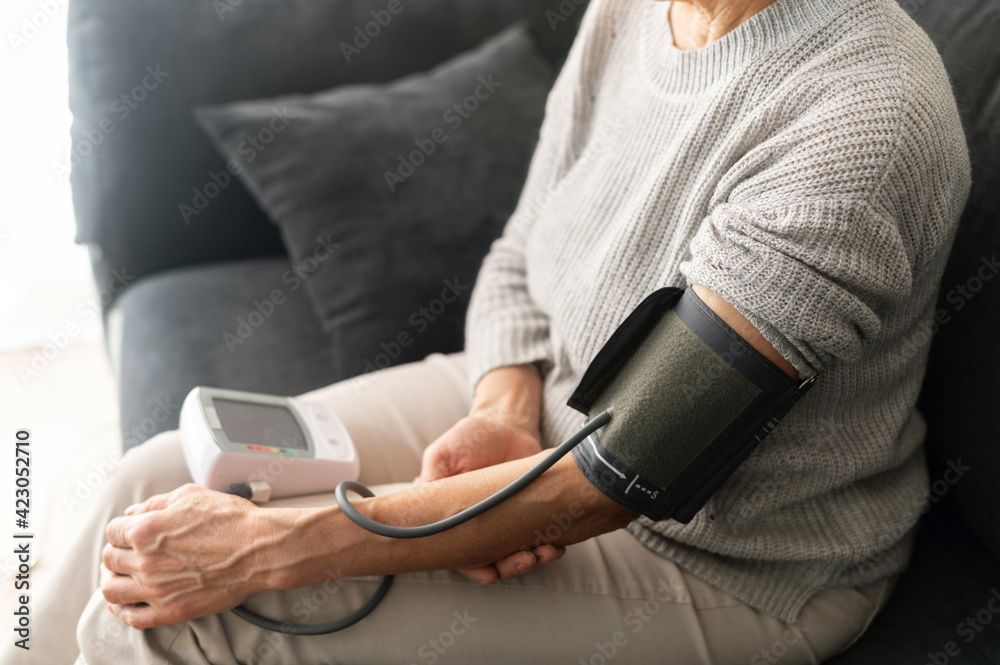 Senior woman checking blood pressure level at home, older female suffering from high blood pressure sitting at a couch and using a pulsometer, tonometer, cropped photo, face is not visible