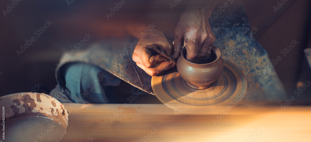 A male artisan in a pottery workshop makes a piece of clay behind a pottery wheel. Traditional pottery craft. - obrazy, fototapety, plakaty 