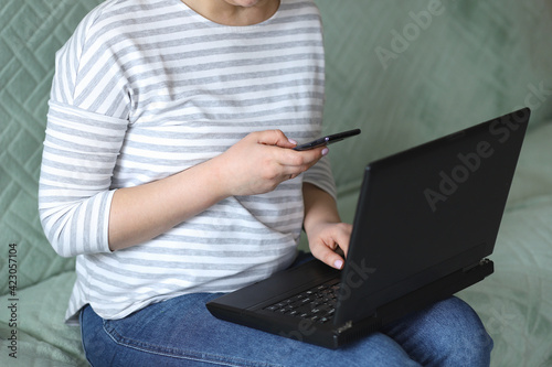 A young woman in a striped sweatshirt and blue jeans sits on the couch at home with a mobile phone in her hands and a laptop and works remotely online. Concept: freelancer. Selective focus. © Tatyana