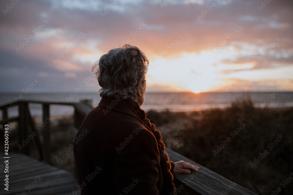Side view of elderly female traveler in casual clothes standing on wooden pier on sandy beach and enjoying seascape at sunset