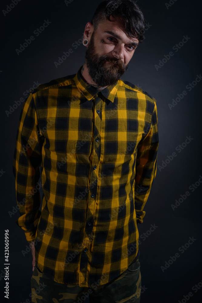 Attractive man with tattoos posing dressed in yellow and black checkered shirt on a black background