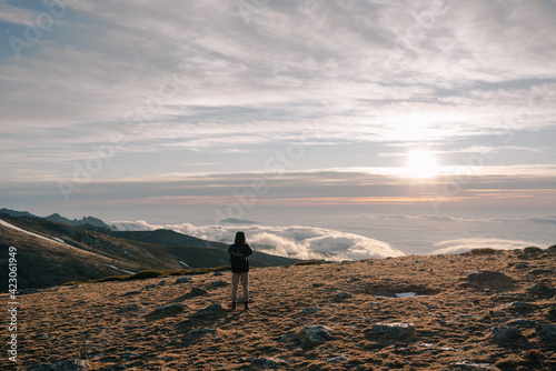 Back view of unrecognizable explorer male tourist in warm outerwear with backpack standing contemplating mountain under thick clouds at dawn