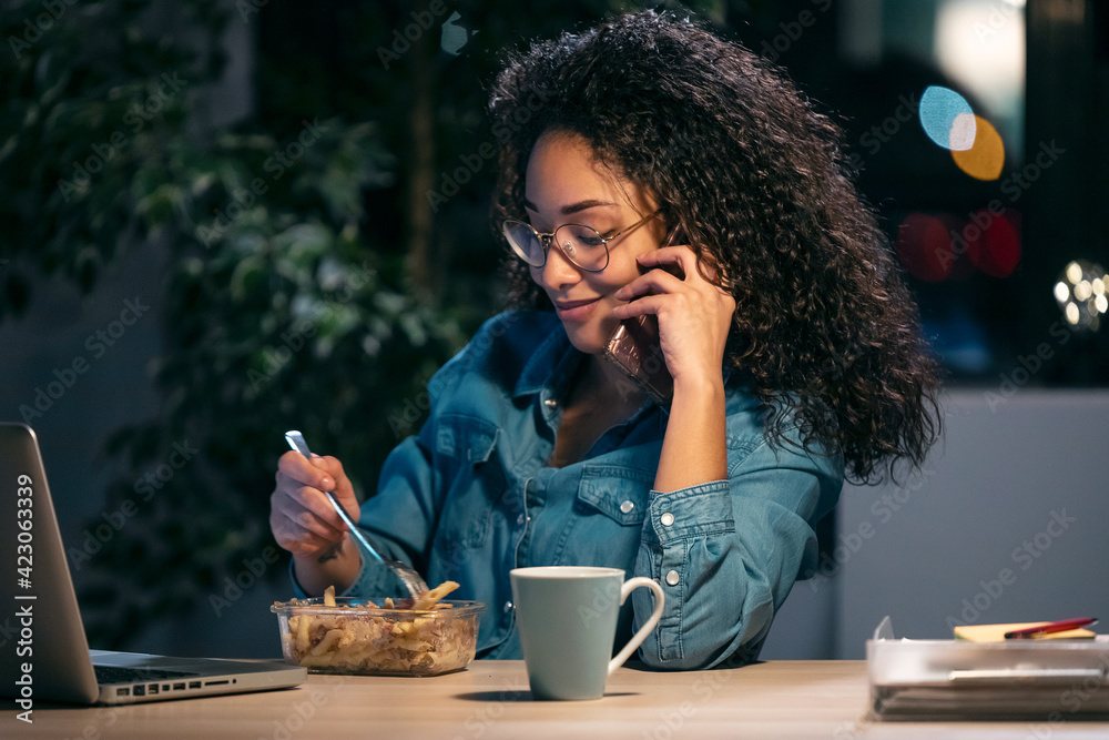 Beautiful afro young business woman working with computer while eating pasta sitting in the office. Beautiful afro young business woman working with computer while eating pasta sitting in the office.