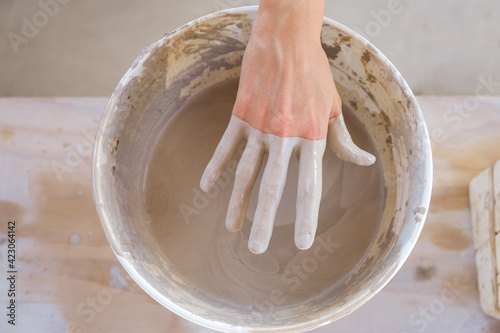 From above crop anonymous craftswoman standing near table and pulling dirty hand from bucket filled with clay in studio in daylight photo