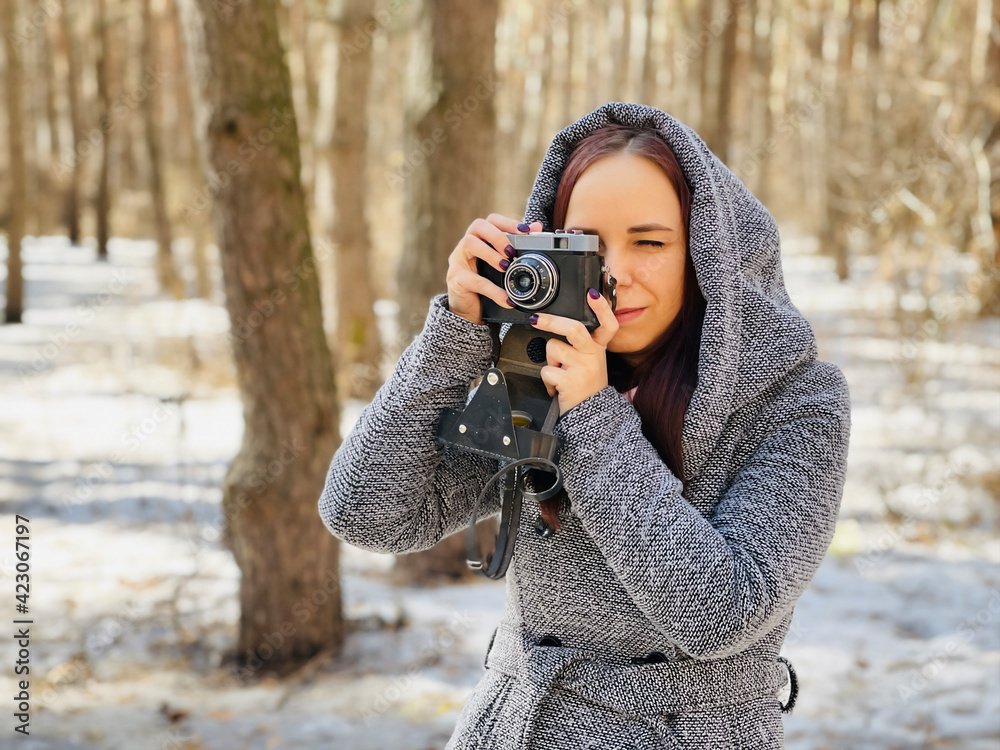 Young woman in gray coat photographing on old photo camera in forest. Pretty female taking photos with old camera in early spring. Young woman in gray coat photographing on old photo camera in forest. Pretty female taking photos with old camera in early spring.