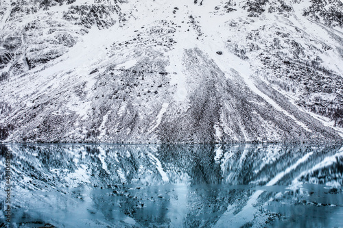 Scenery of mountain slope covered with snow and leafless trees reflecting in smooth water surface of lake in winter in Norway