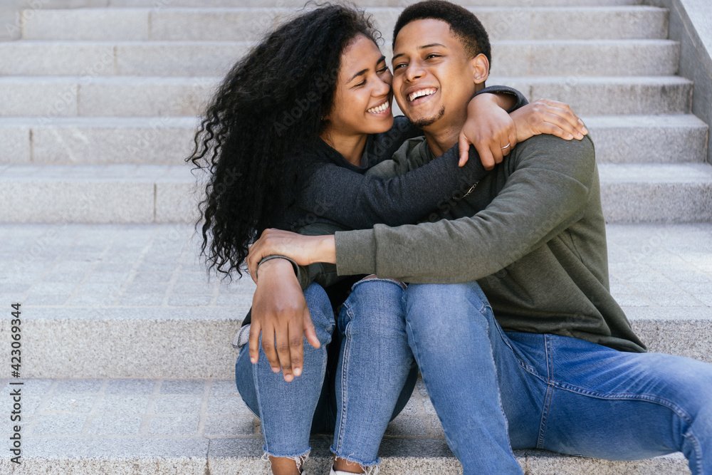 happy interracial couple hugging and smiling at each other sitting on the stairs. Young African American friends enjoying themselves outside the building. The curly haired afro girl 