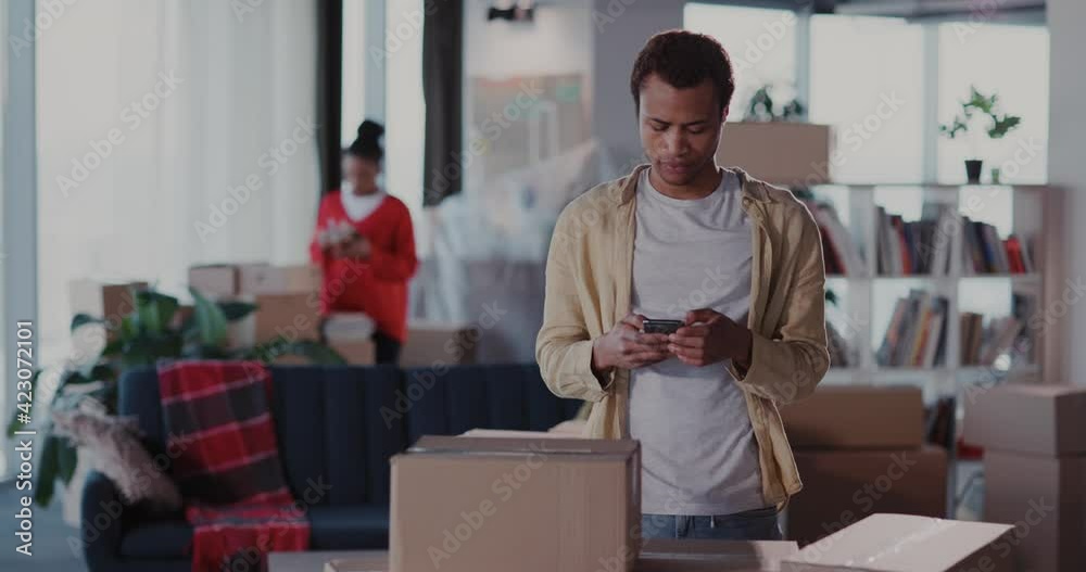 Black handsome young man moving house ordering taxi on mobile phone. His busy young wife collecting personal items into carton boxes. Moving out. Family. Black handsome young man moving house ordering taxi on mobile phone. His busy young wife collecting personal items into carton boxes. Moving out. Family.