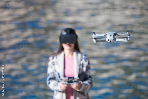 Excited female wearing modern VR goggles operating drone with remote controller and experiencing virtual reality while standing against blurred sea