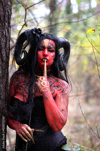 Sexy Teufelin steht im Wald. Sie hat die Hörner auf dem Kopf, eine schwarze Corsage. sie ist mit der roten Farbe und der Muster angemalt.Sie hat lange, schwarze Haare. Nah Portrait.