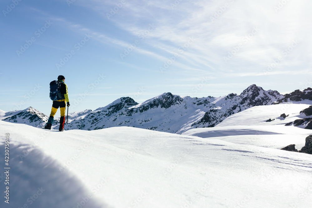 Full body back view of unrecognizable male mountaineer in warm activewear with backpack standing on slope of snowy rocky mountain and enjoying spectacular landscape in sunny winter day Full body back view of unrecognizable male mountaineer in warm activewear with backpack standing on slope of snowy rocky mountain and enjoying spectacular landscape in sunny winter day