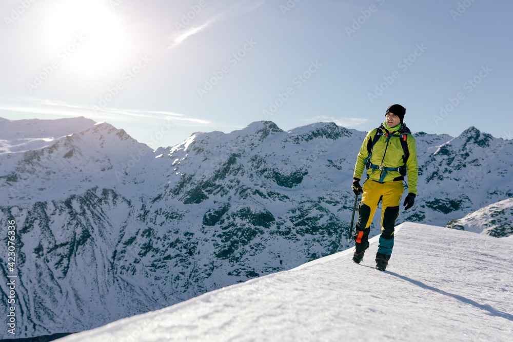 Full body male mountaineer in warm activewear with backpack standing on slope of snowy rocky mountain and enjoying spectacular landscape in sunny winter day Full body male mountaineer in warm activewear with backpack standing on slope of snowy rocky mountain and enjoying spectacular landscape in sunny winter day