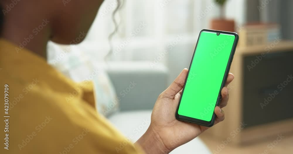 Close up of unrecognizable African American woman using smartphone with green screen and scrolling while sitting in room. Female holding black mobile phone with chroma key in hand, tech concept