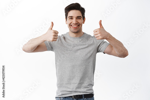 Portrait of cheerful man in basic clothing smiling and showing thumbs up at camera isolated over white background