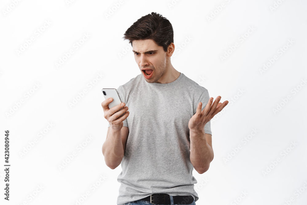 Frustrated man look confused at smartphone screen, read strange message on mobile phone and arguing, complaining, standing against white background Frustrated man look confused at smartphone screen, read strange message on mobile phone and arguing, complaining, standing against white background