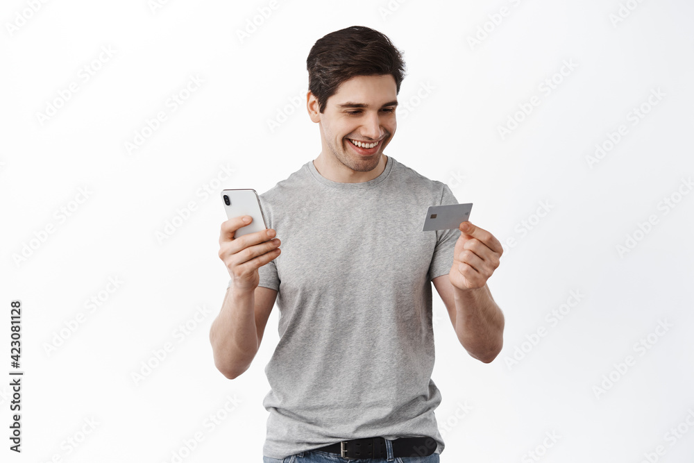Handsome man paying online with credit card and smartphone, shopping in app, order something in internet shop, standing over white background Handsome man paying online with credit card and smartphone, shopping in app, order something in internet shop, standing over white background