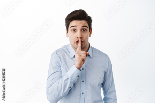 Corporate man in shirt asking to keep quiet, gossiping in office, shushing, holding finger to lips and looking excited, white background