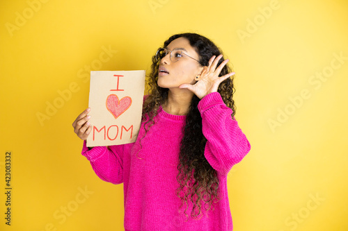 Beautiful woman celebrating mothers day holding poster love mom message surprised with hand over ear listening an hearing to rumor or gossip