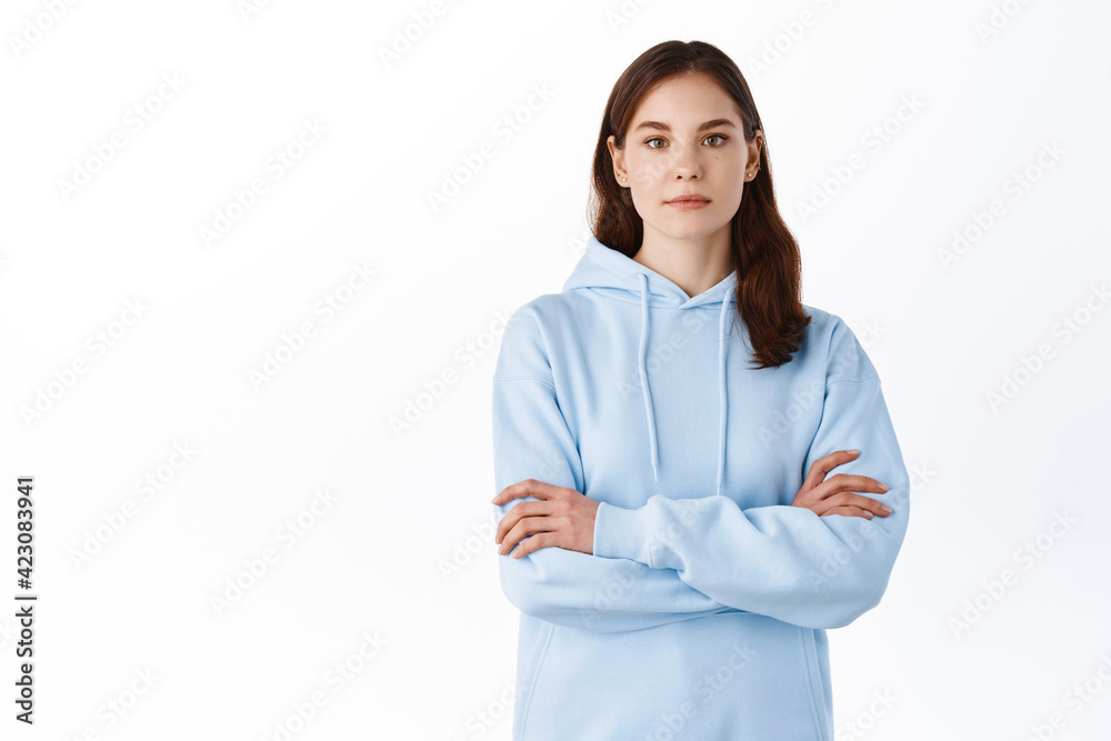 Serious-looking girl student in hoodie, cross arms on chest and look like professional with determined face, standing against white background Serious-looking girl student in hoodie, cross arms on chest and look like professional with determined face, standing against white background