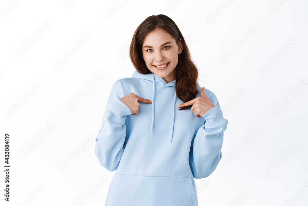Cute brunette girl in casual hoodie pointing at herself, smiling and self-promoting, choose me gesture, standing against white background Cute brunette girl in casual hoodie pointing at herself, smiling and self-promoting, choose me gesture, standing against white background
