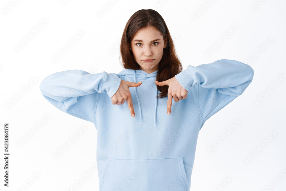 Young upset girl with gloomy sad face, pointing fingers down at bottom copy space, complaining about unfair situation, standing displeased against white background Young upset girl with gloomy sad face, pointing fingers down at bottom copy space, complaining about unfair situation, standing displeased against white background