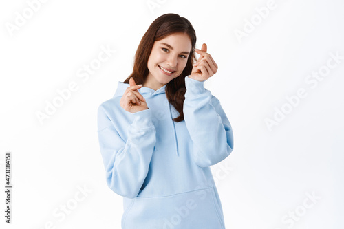 Attractive female model in hoodie showing finger hearts gesture and smiling cute, express love and tenderness, standing against white background