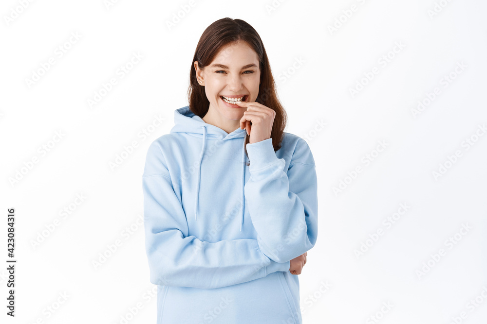 Portrait of positive and cute young woman in hoodie, smiling and laughing, touching lips with cheerful white smile, standing against studio background