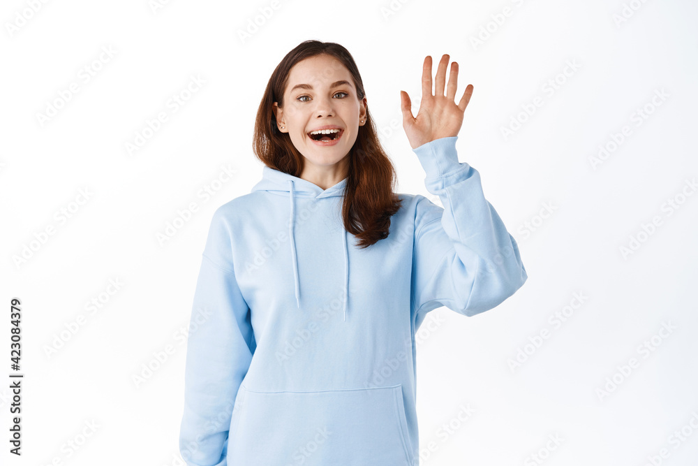Friendly girl student saying hello and waving hand. Cheerful woman model greeting or saying goodbye, making hi gesture, standing against white background Friendly girl student saying hello and waving hand. Cheerful woman model greeting or saying goodbye, making hi gesture, standing against white background