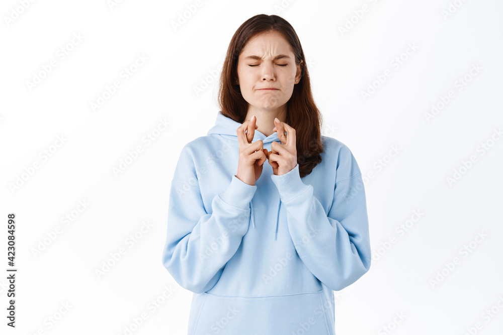 Young woman praying with intense face, put effort in pleading god for dream to come true, close eyes and cross fingers for good luck, making wish, standing against white background Young woman praying with intense face, put effort in pleading god for dream to come true, close eyes and cross fingers for good luck, making wish, standing against white background
