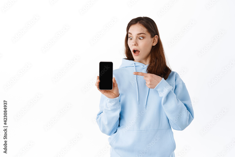Shocked young woman pointing finger at her phone screen, gasping amazed while staring at smartphone display, standing against white background